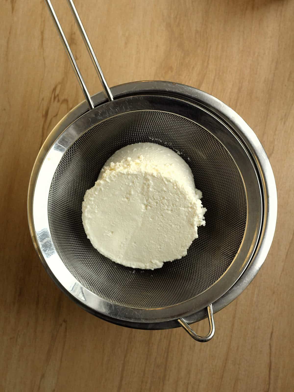 Ricotta placed in a fine mesh sieve over a bowl for draining to make ricotta fritters.
