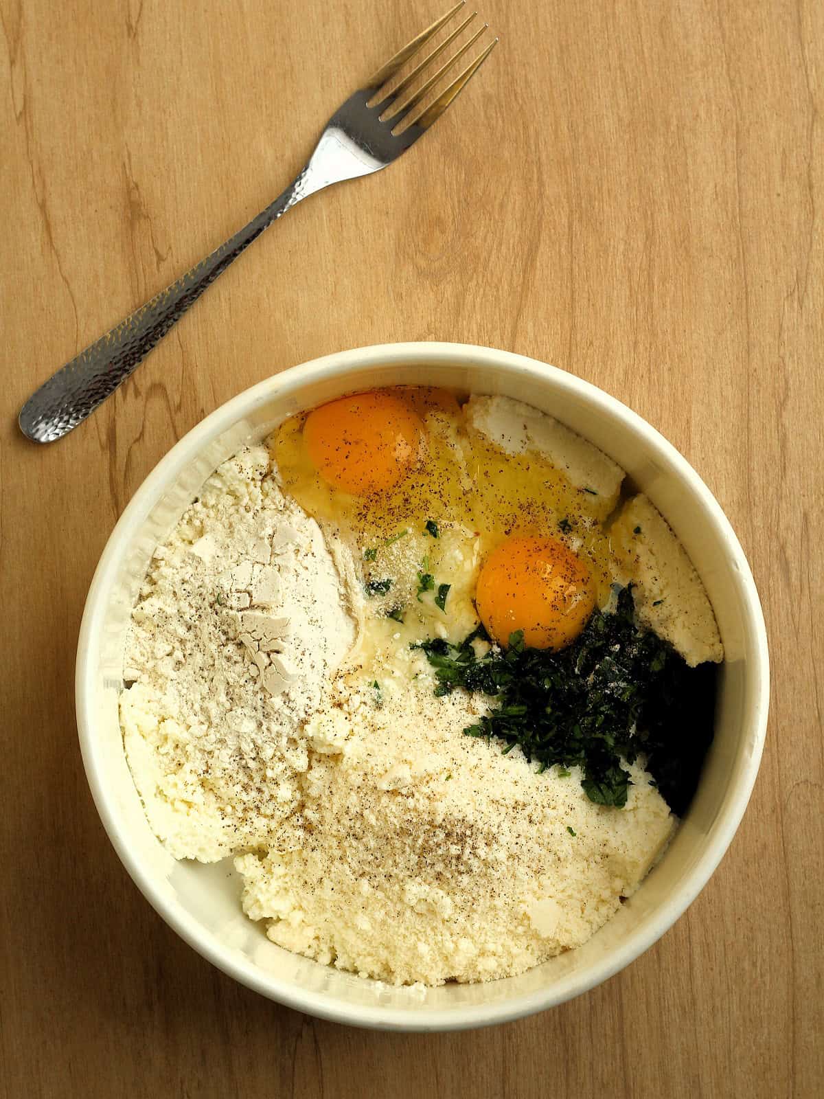 Ingredients for savoury ricotta cheese fritters added to bowl next to fork.