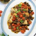Sicilian swordfish with cherry tomatoes, olives and capers plated with parsley sprig on side.