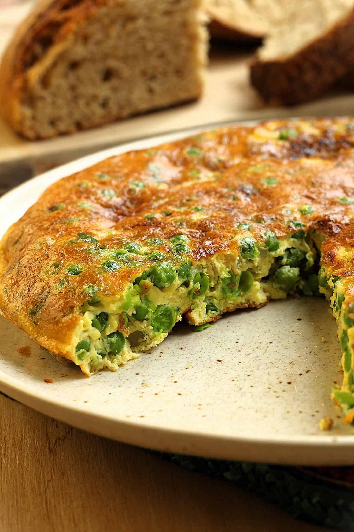 Frittata with peas with a slice cut out on plate next to loaf of bread.