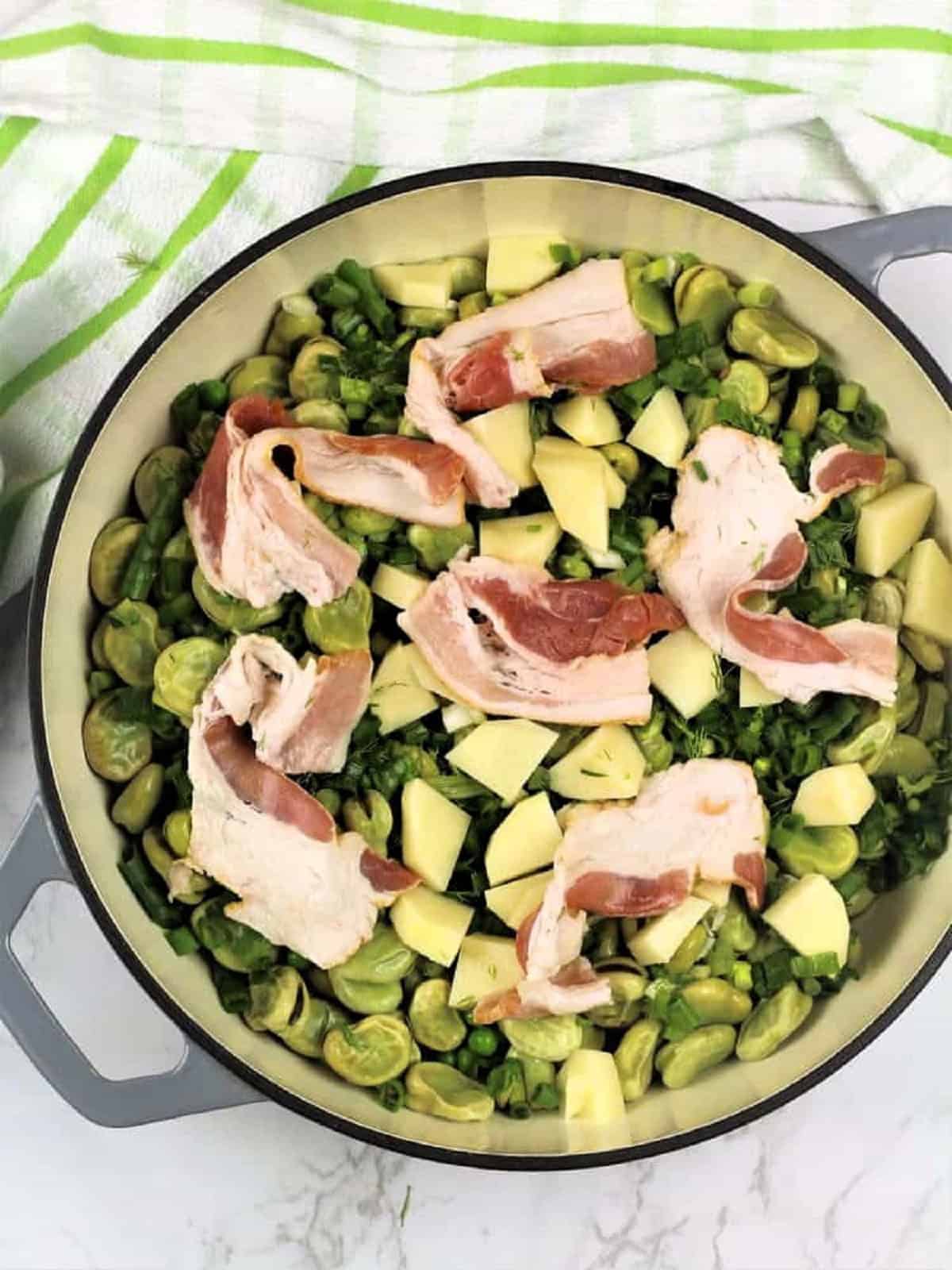 Large skillet filled with fava beans, peas, potato, green onions, fennel fronds and bacon slices.