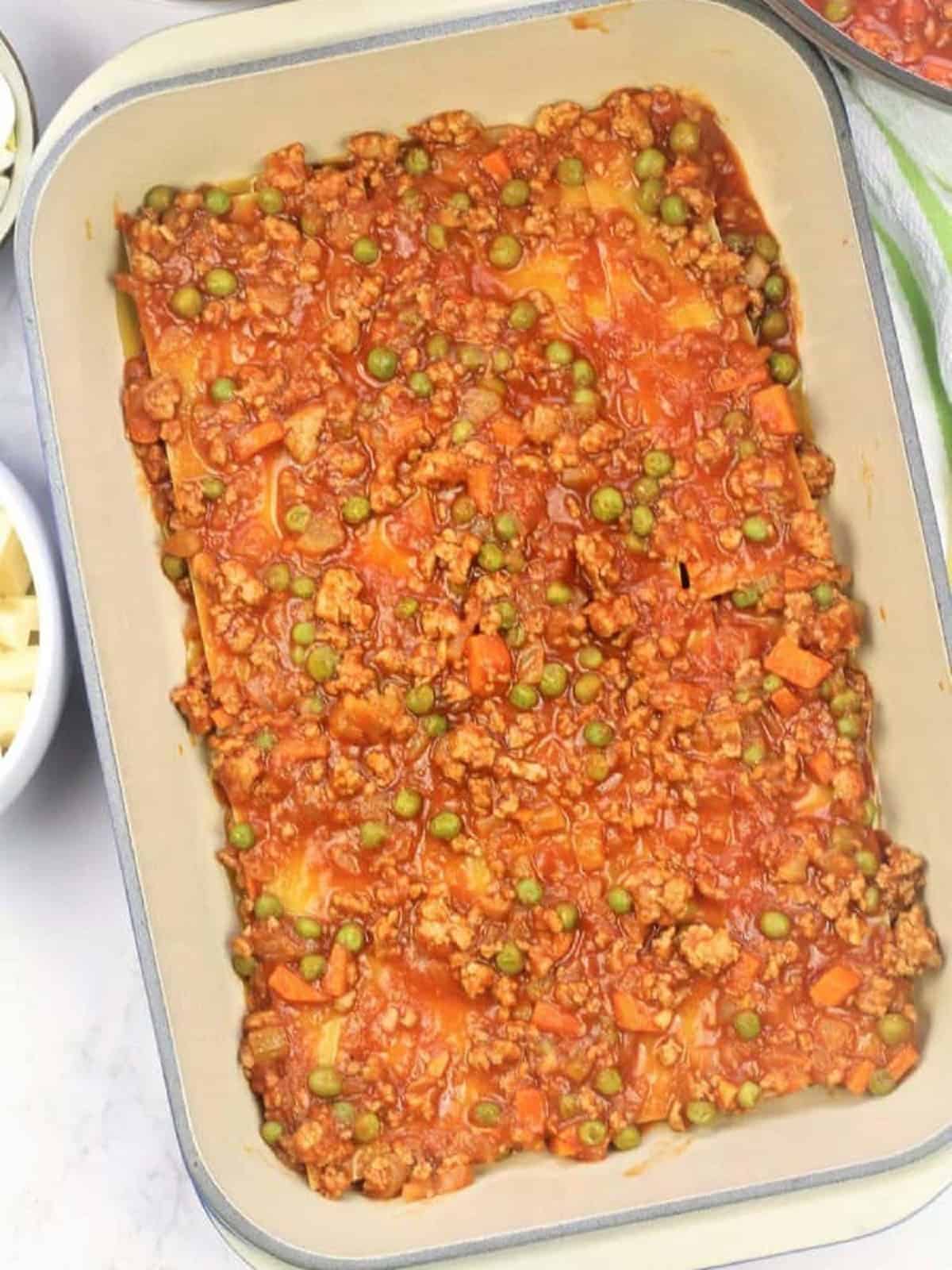 Lasagna sheets covered with meat sauce with peas in casserole dish.