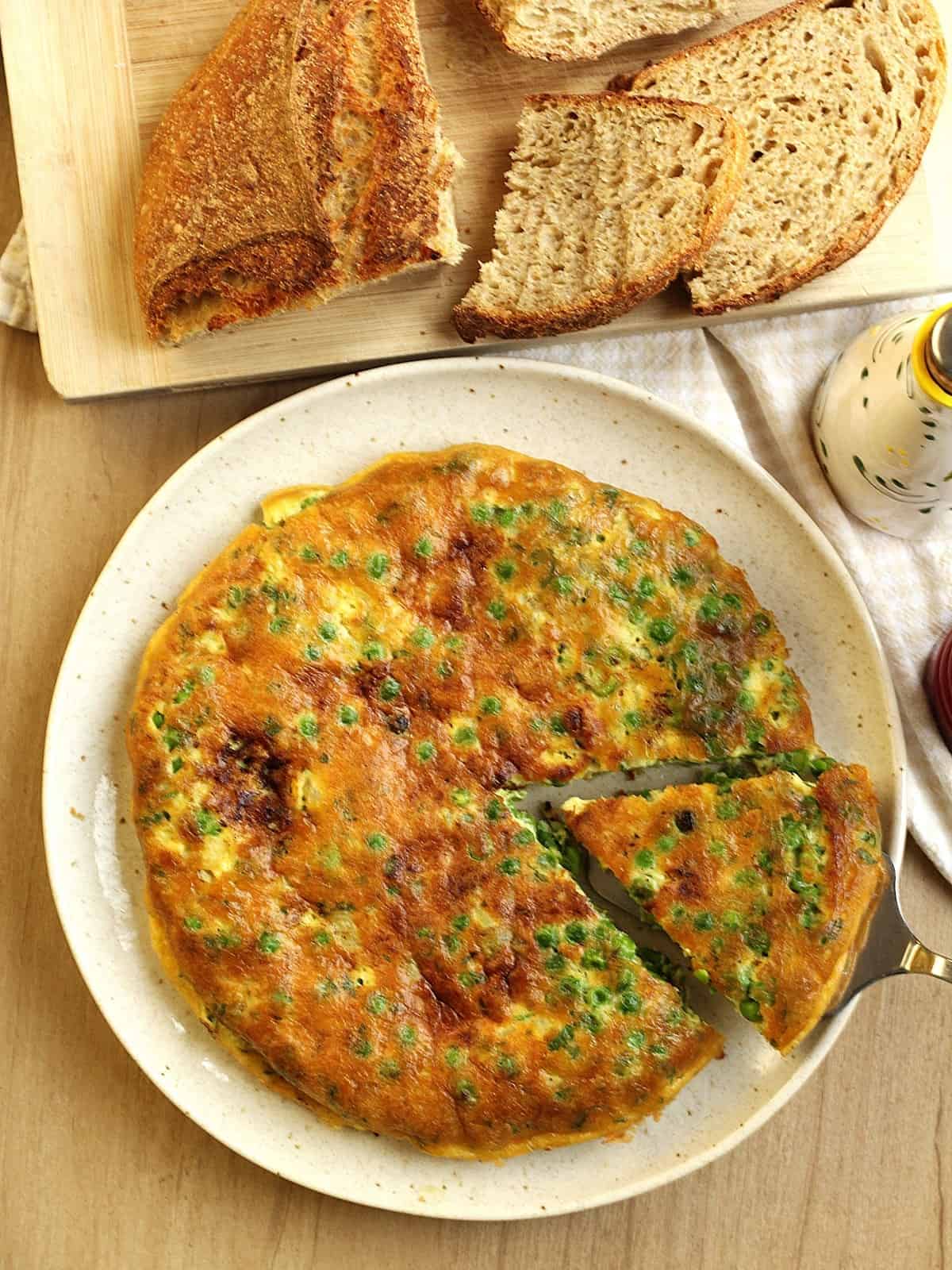 Frittata with peas with spatula serving a wedge next to wood board with sliced bread.
