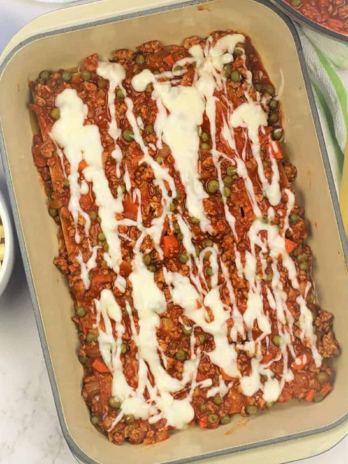Béchamel sauce drizzled over lasagna with meat sauce in casserole dish.