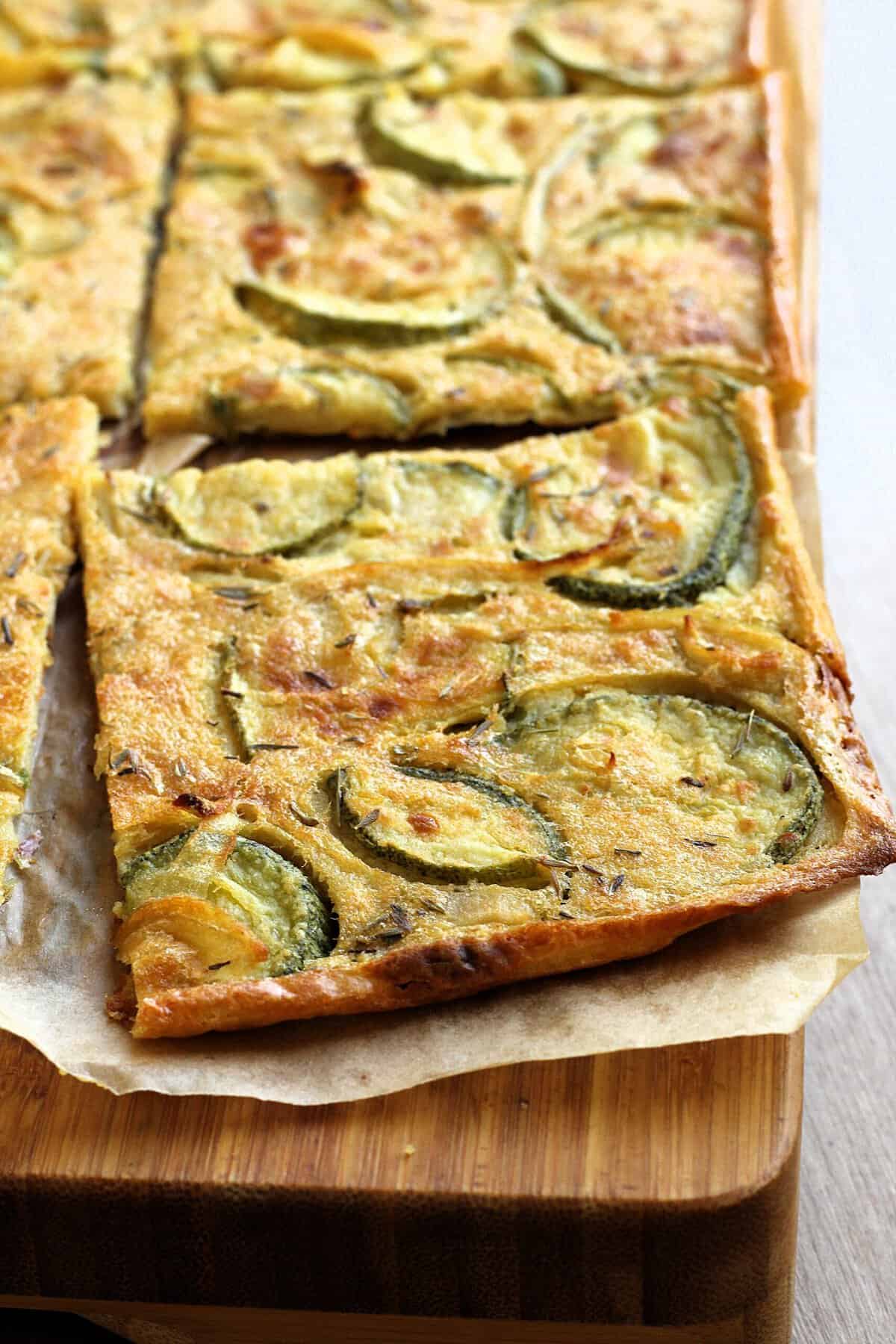 Square of chickpea flatbread with zucchini and onions on wood board.