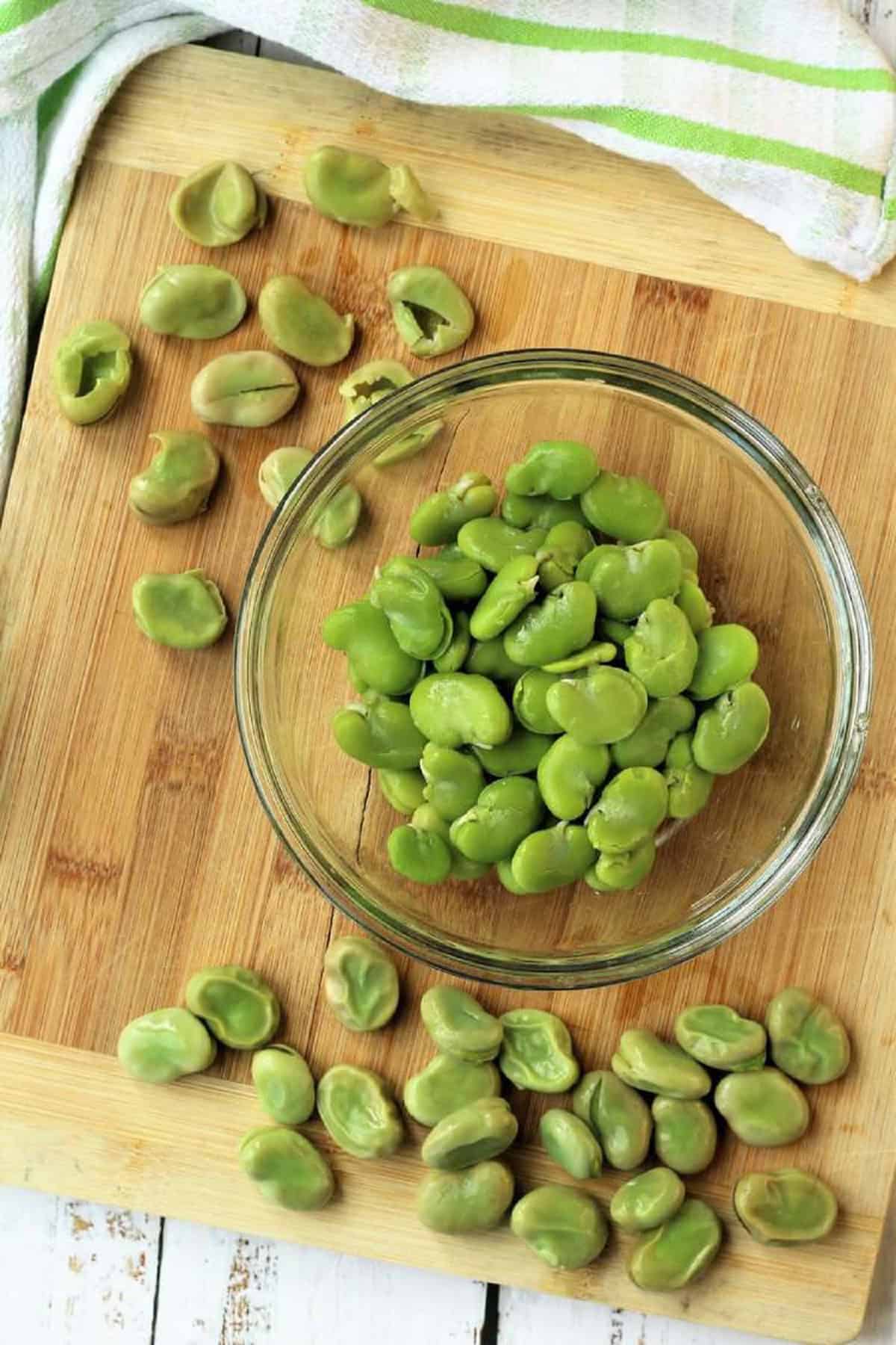 Fava beans in bowl with skin peeled off on wood board.