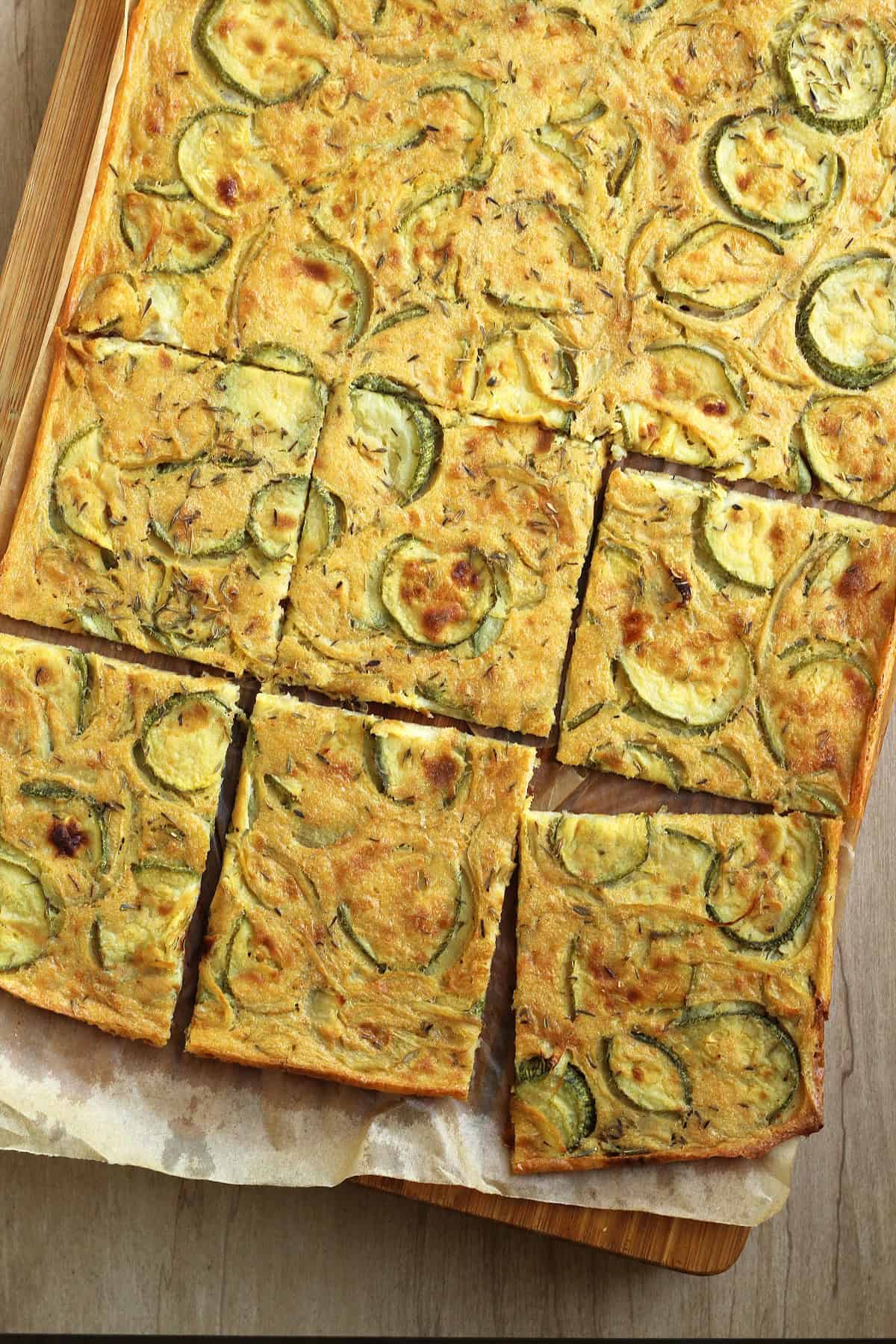Baked chickpea flour flatbread with zucchini cut into squares on wood board.