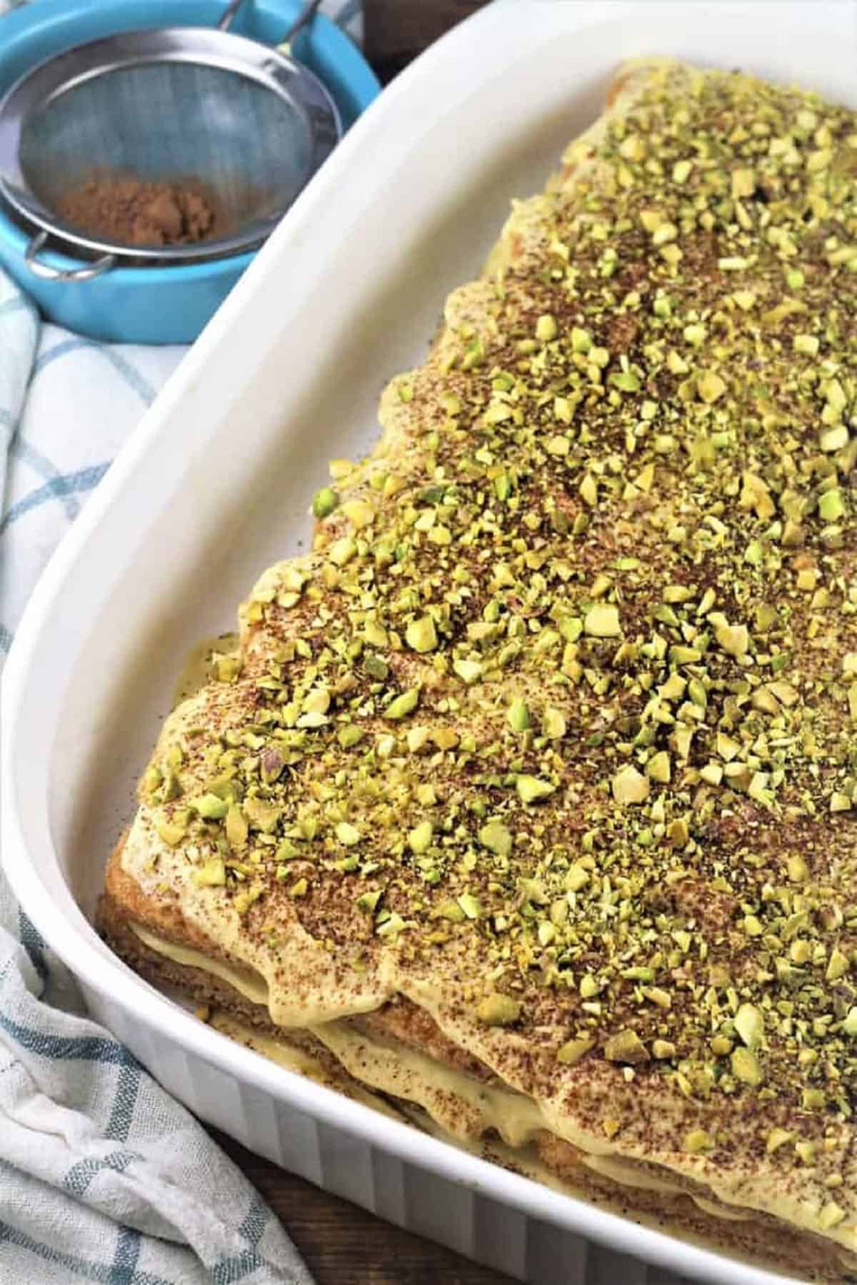 Tiramisu with pistachio cream in white casserole dish topped with chopped pistachios.
