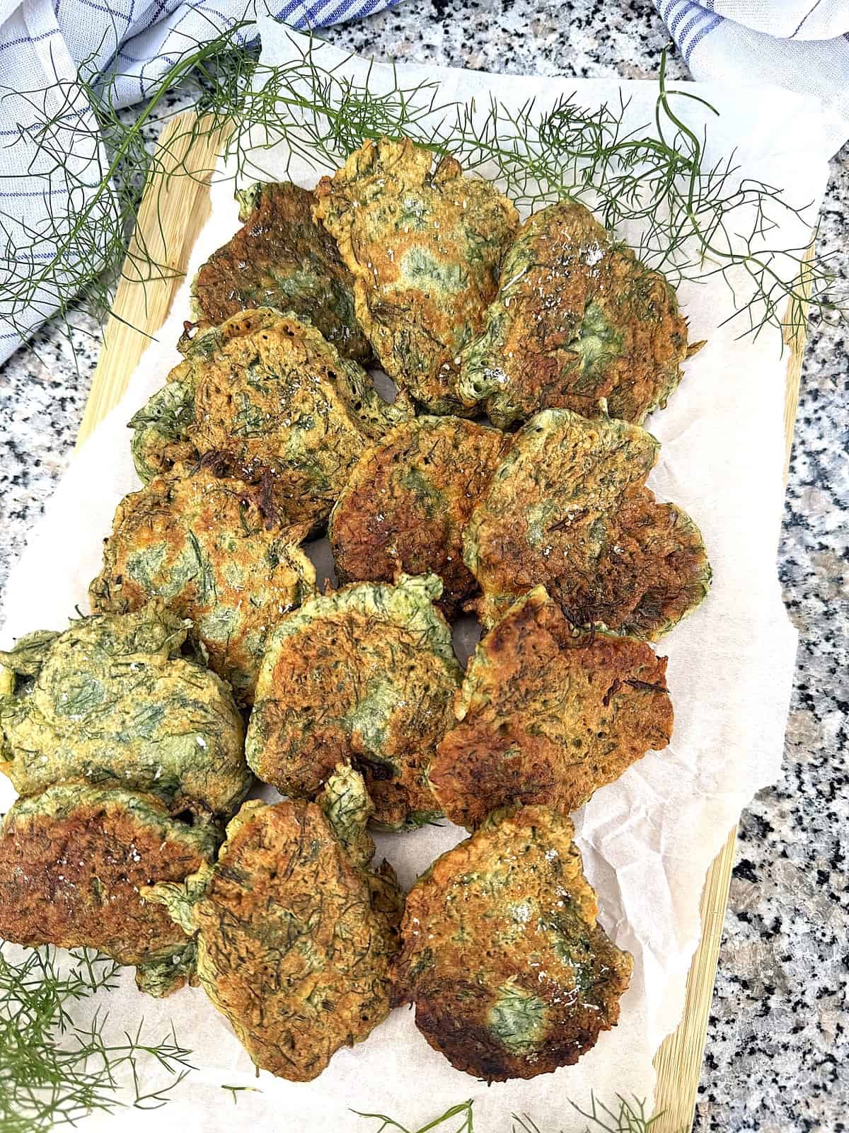 Sicilian wild fritters (frittelle di finocchietto) layered on wood board with fennel fronds around them.