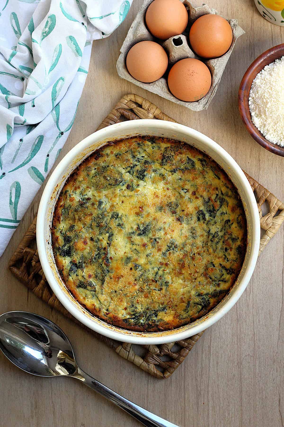 Tortino di bietole e ricotta in round casserole dish surrounded by eggs, bowl of cheese and serving spoon.